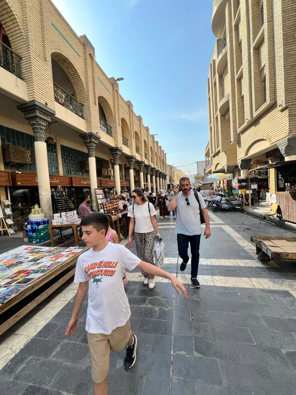 Mutanabbi Street in Baghdad in the afternoon, colonnaded brick arcades lining the book market as people walk the paved street.