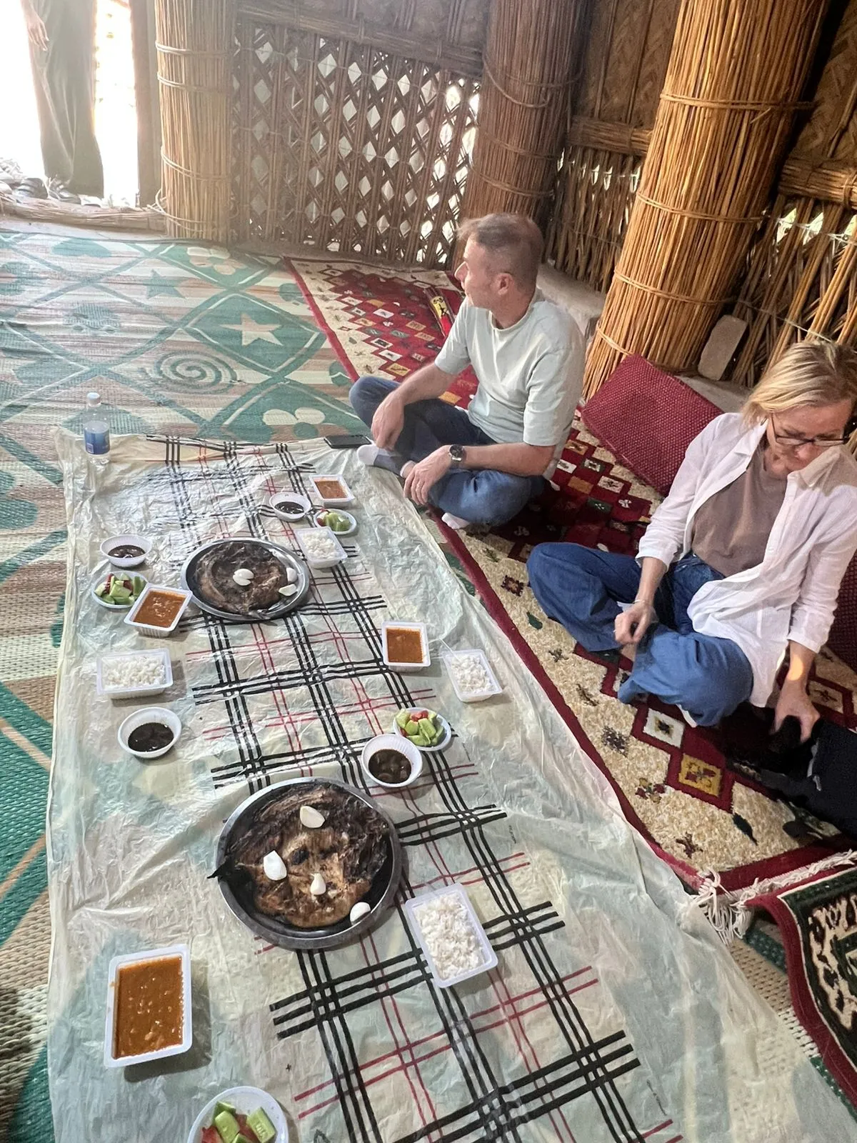 A meal laid out on a woven mat inside a traditional reed mudhif in the Mesopotamian Marshes, arched bundles of qasab reed rising overhead.